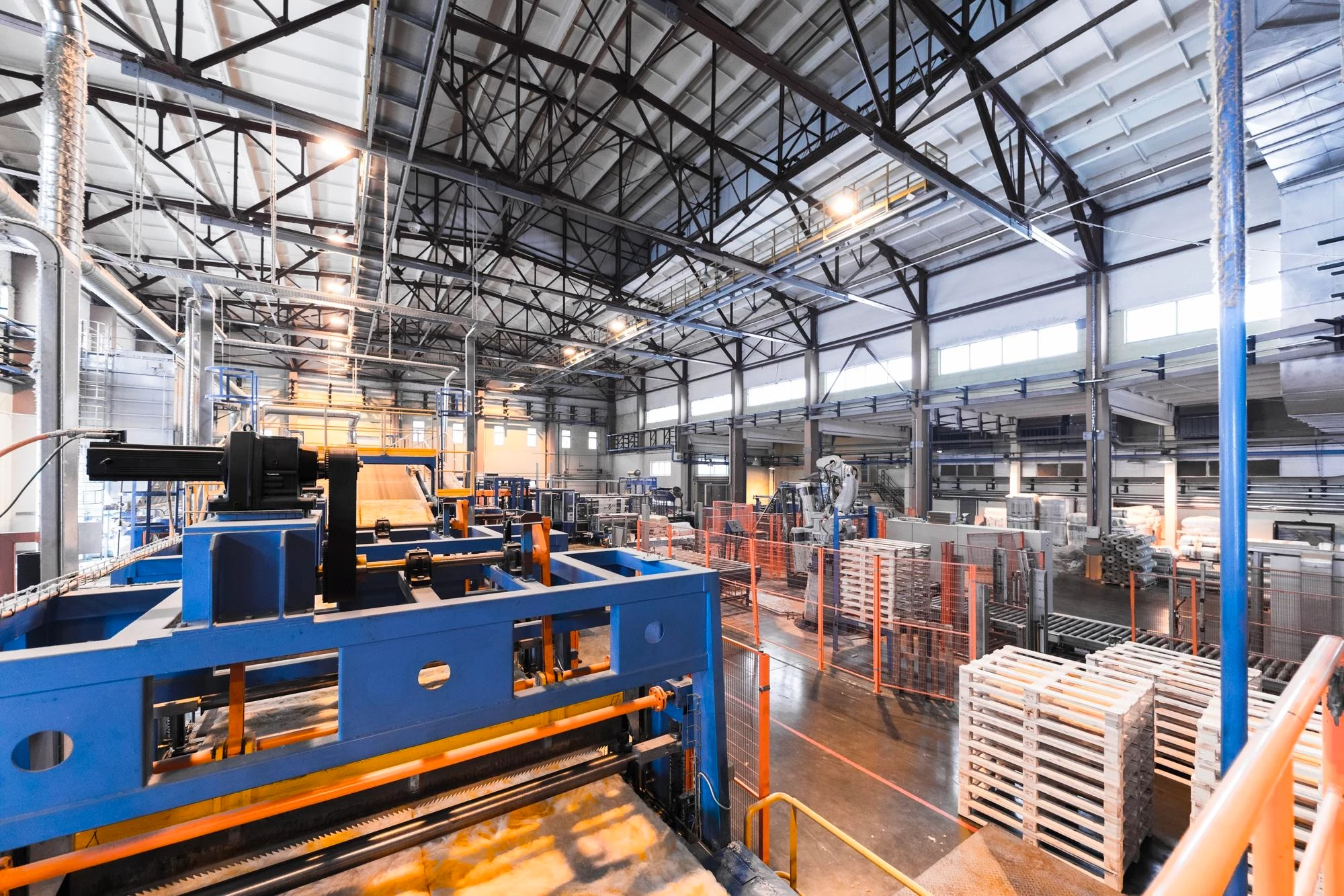 A-wide-shot-of-a-brightly-lit,-industrial-factory-interior,-showcasing-large-blue-machinery-in-the-foreground-and-stacks-of-pallets-in-the-background-under-a-complex-network-of-ceiling-beams-and-pipes. A wide shot of a brightly lit, industrial factory interior, showcasing large blue machinery in the foreground and stacks of pallets in the background under a complex network of ceiling beams and pipes.