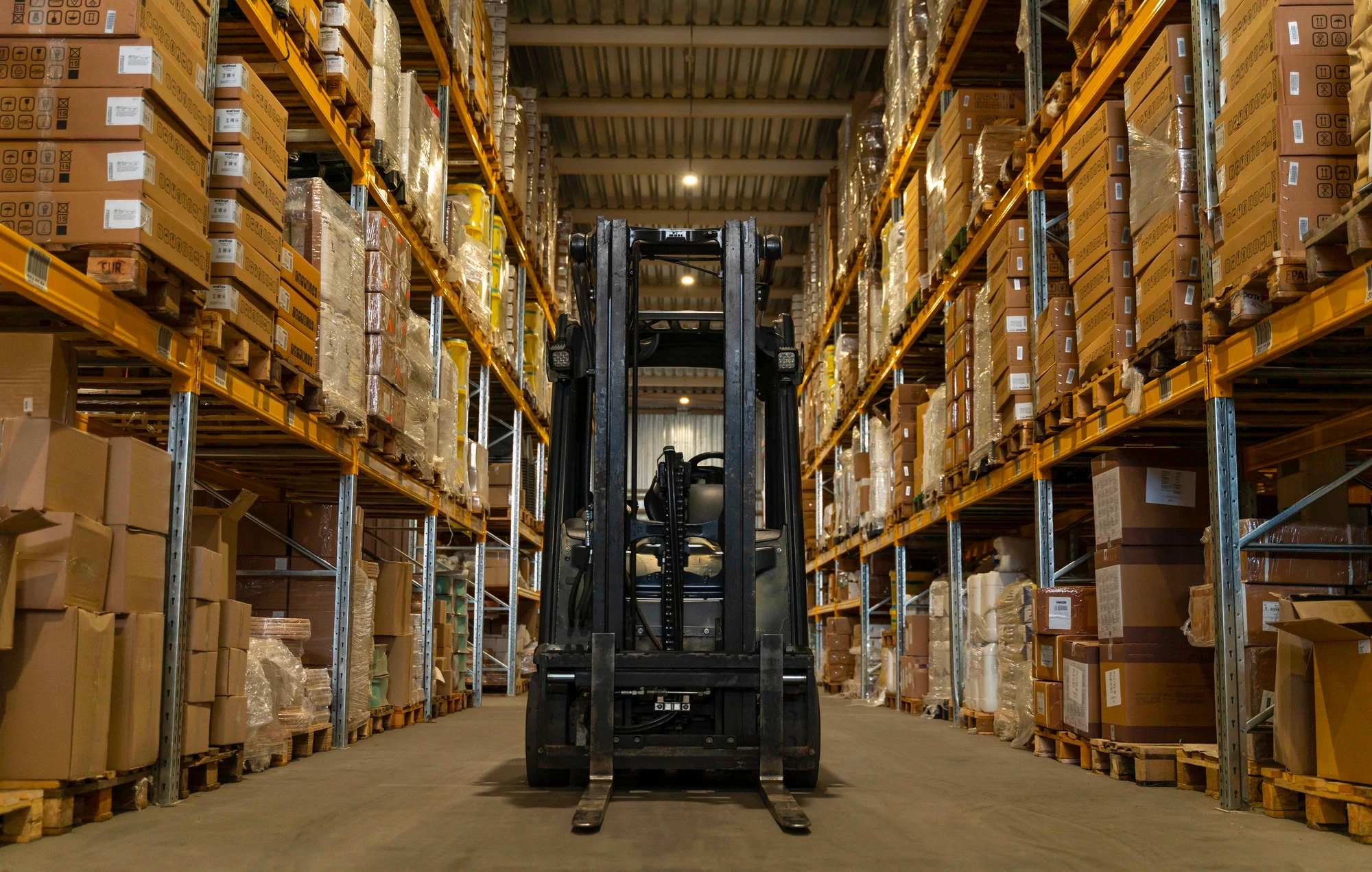 A-forklift-sits-centered-in-the-aisle-of-a-warehouse,-surrounded-by-tall-racks-filled-with-boxes-and-packaged-goods. A forklift sits centered in the aisle of a warehouse, surrounded by tall racks filled with boxes and packaged goods.