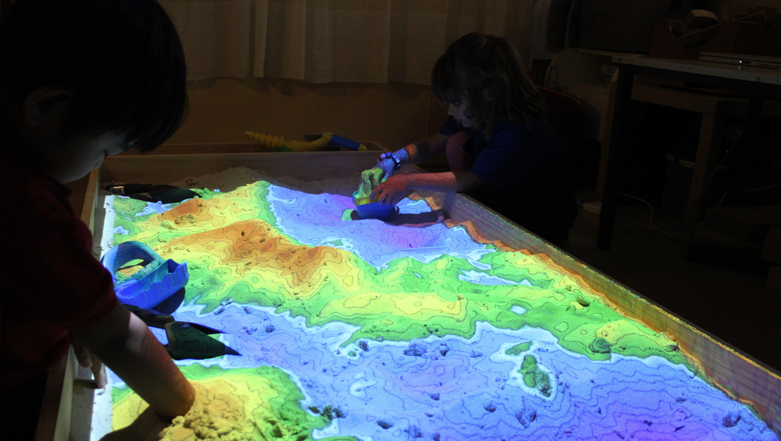 Children playing with a topographic sandbox, projecting elevation color mapping onto the sand.