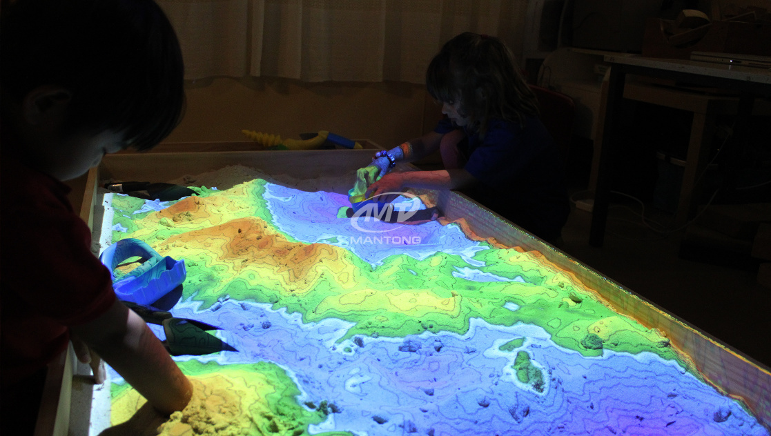 Children playing with a topographic sandbox, projecting elevation color mapping onto the sand. Children playing with a topographic sandbox, projecting elevation color mapping onto the sand.