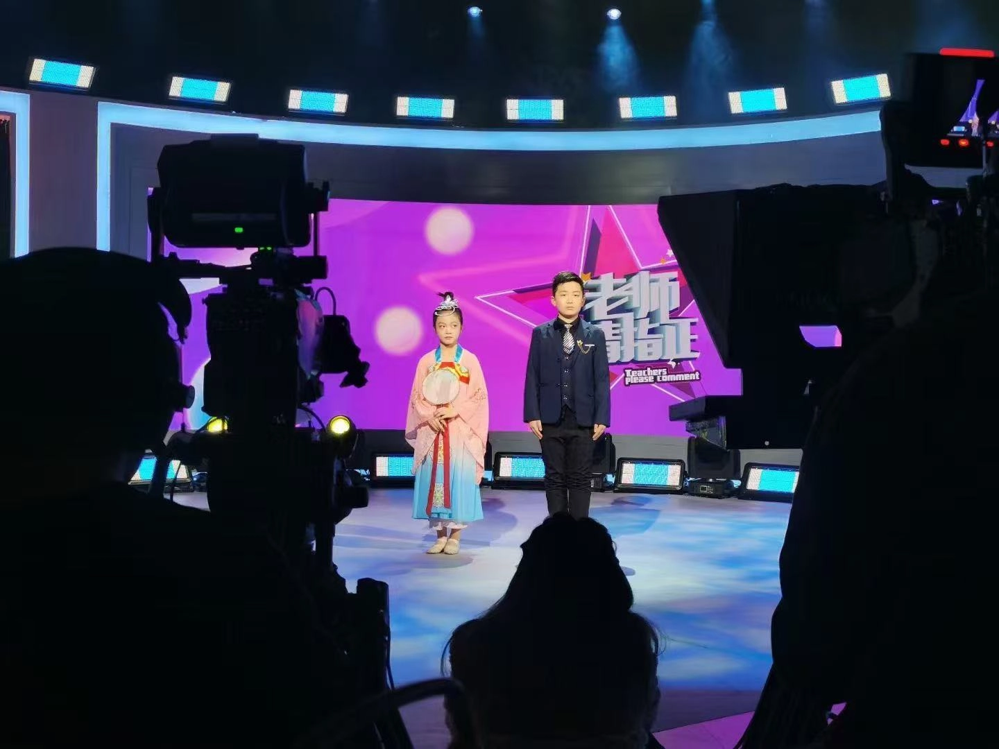 Two children on stage, one in traditional Chinese dress, filmed by cameras. - Vanray