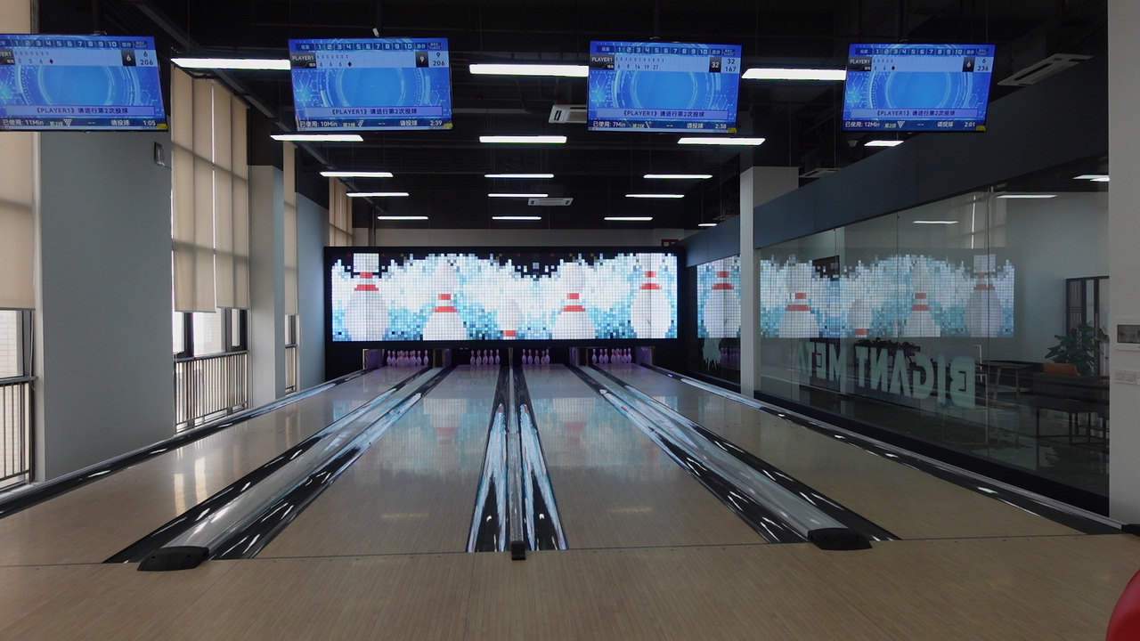 An image of a bowling alley with four lanes. The lanes are made of wood and have a shiny, waxed surface. The pins are set up at the end of each lane. There is a large screen at the end of the alley that shows the scores of the bowlers.