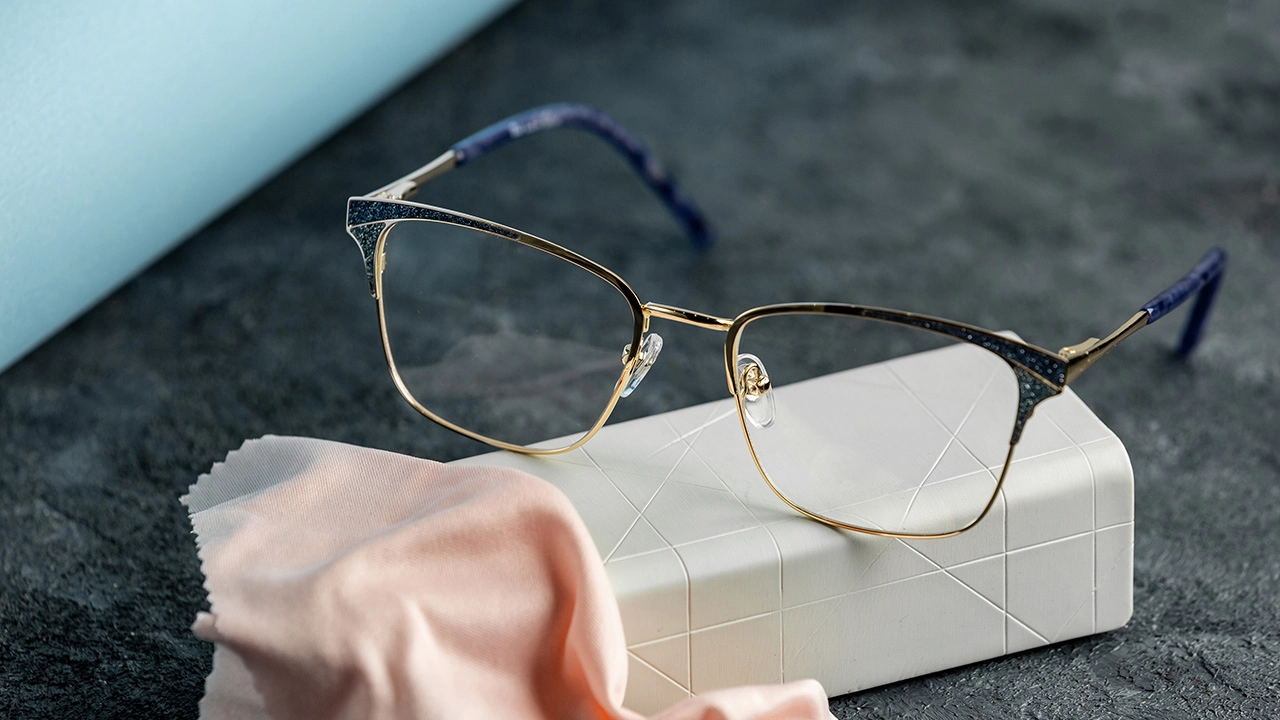 Gold-framed eyeglasses with blue glitter accents on a white case with a pink cleaning cloth. - KARUSON