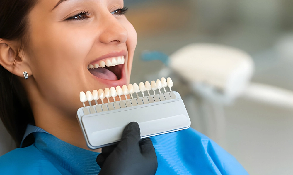 A woman is sitting in a dentist's chair with her mouth open while the dentist holds a shade guide to her teeth.