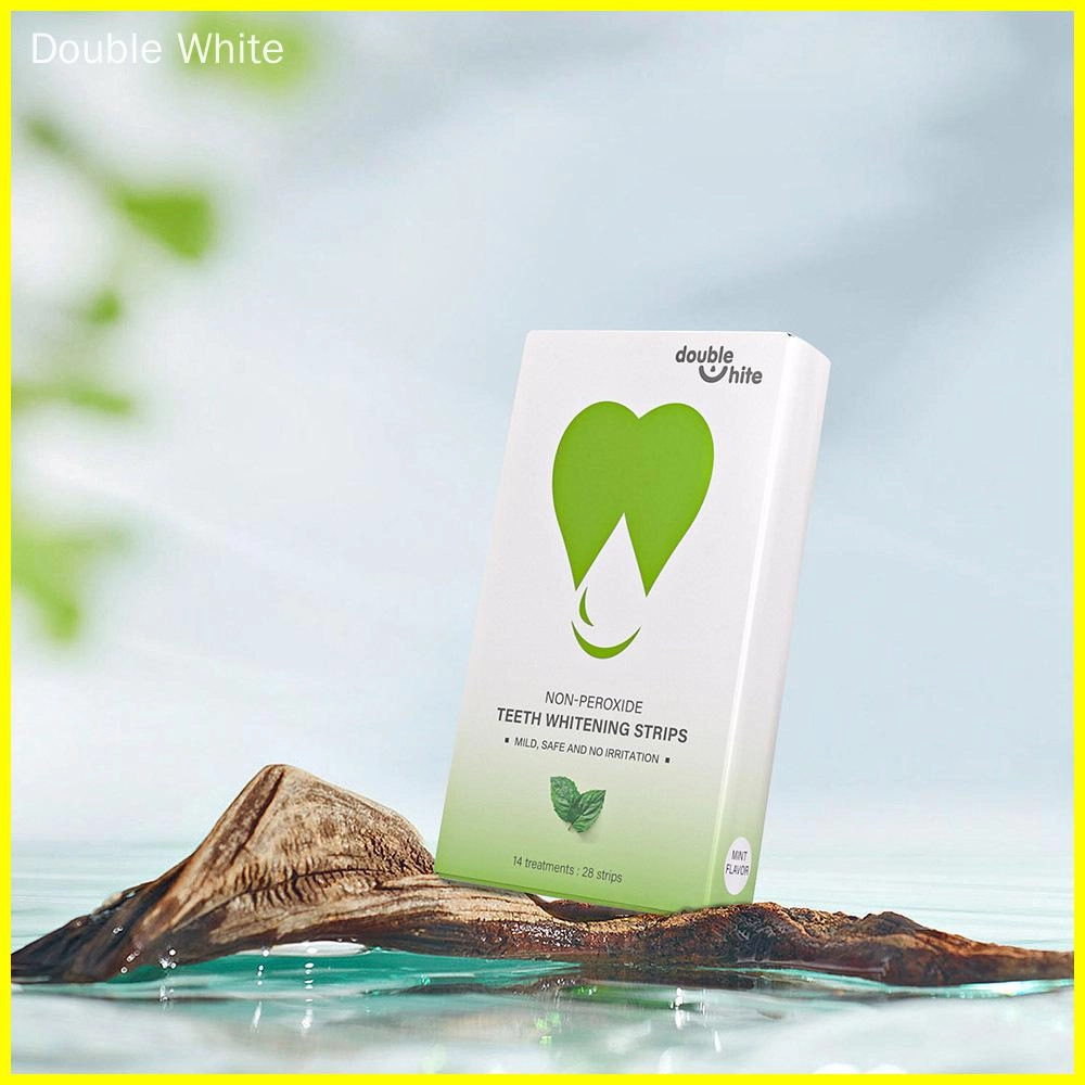 Коробка полосок для отбеливания зубов Double White без перекиси. Белая коробка с изображением зелёного листа на лицевой стороне. Коробка стоит на деревянной доске в луже воды. - Double White
