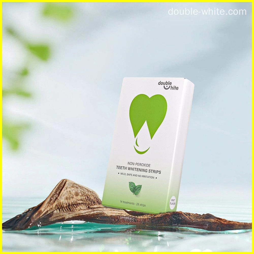 Uma caixa de tiras de clareamento dental sem peróxido Double White. A caixa é branca com um desenho de folha verde na frente. A caixa está sobre um pedaço de madeira em uma poça d'água. - Double White