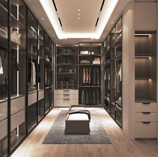 Modern luxury walk-in closet with glass doors, built-in lighting, and an ottoman.