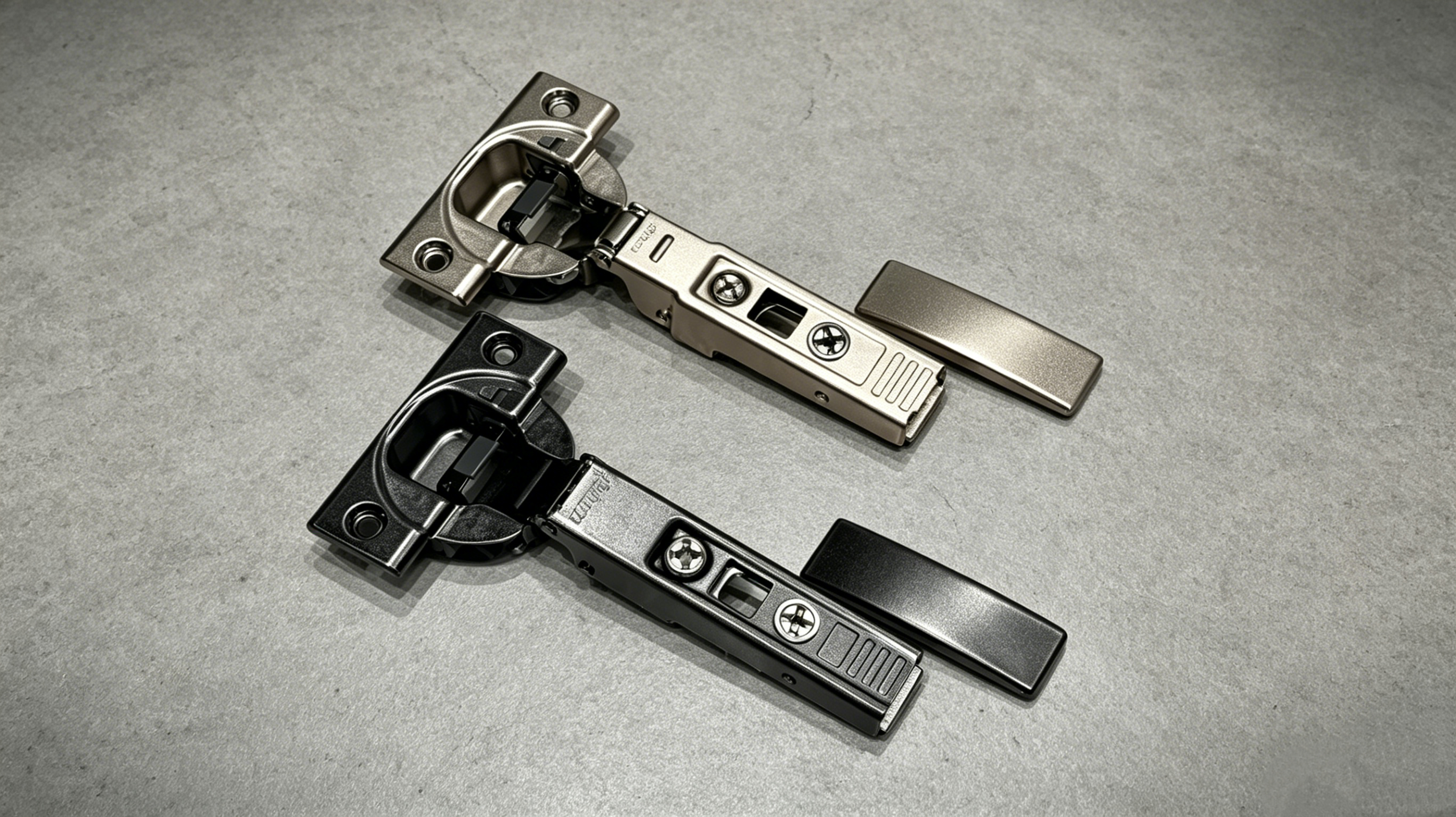 Two concealed cabinet hinges in nickel and black finishes on a gray surface.