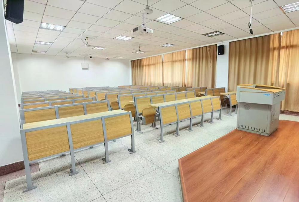 Empty lecture hall with light wood desks and chairs, a podium, and projector.