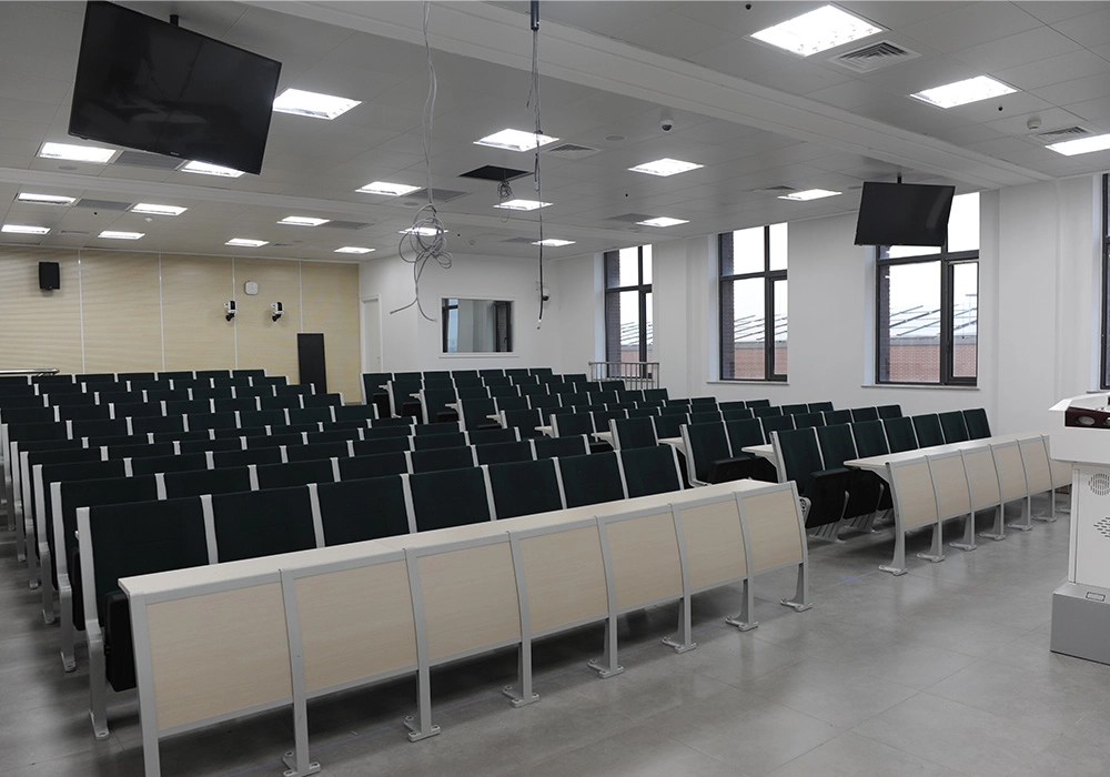 Empty university lecture hall with dark-colored seats and light-colored desks, two flat-screen TVs mounted on the walls, and large windows.
