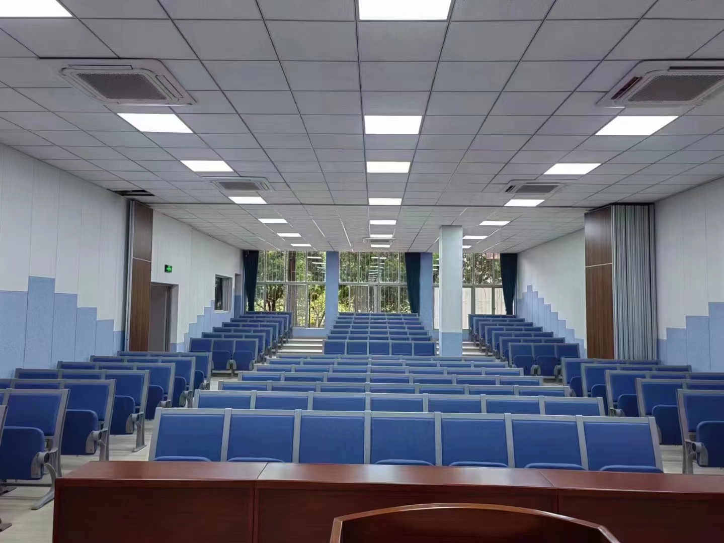 Empty lecture hall with blue seating and a view of trees outside.