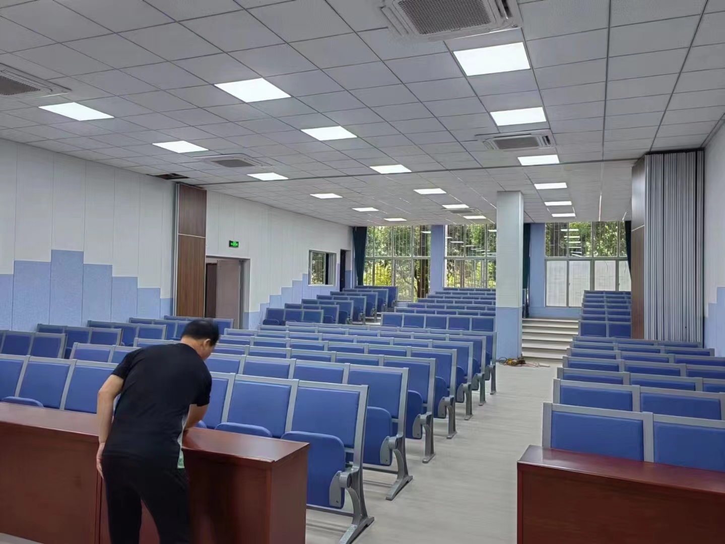 Worker cleaning desks in a large lecture hall with blue seating and tiered rows.