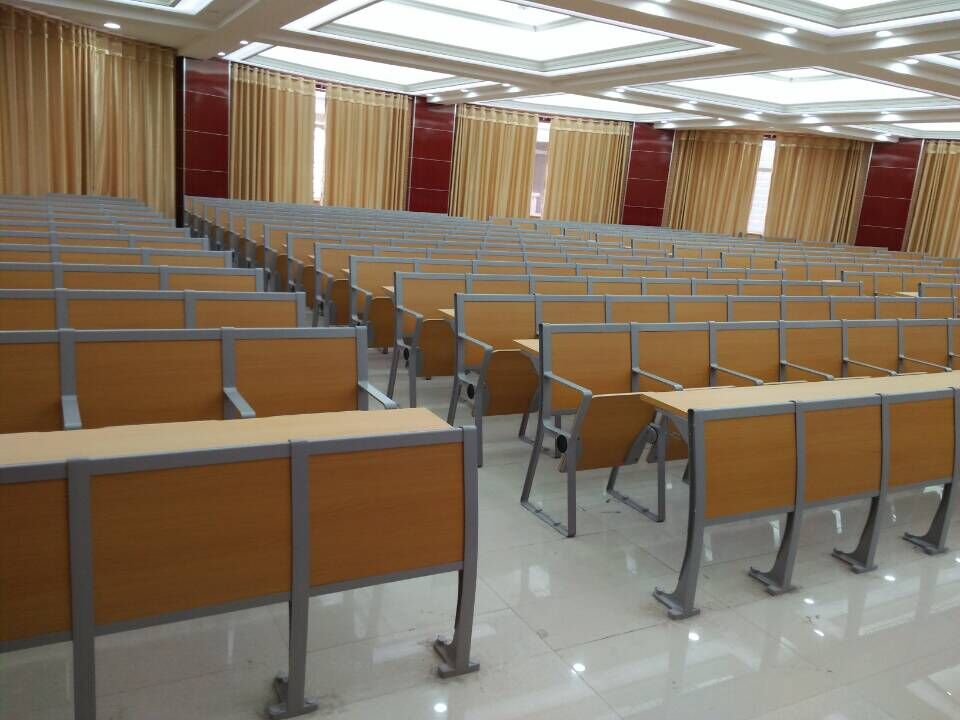 Rows of light brown classroom desks and chairs in a large lecture hall with beige curtains. Rows of light brown classroom desks and chairs in a large lecture hall with beige curtains.