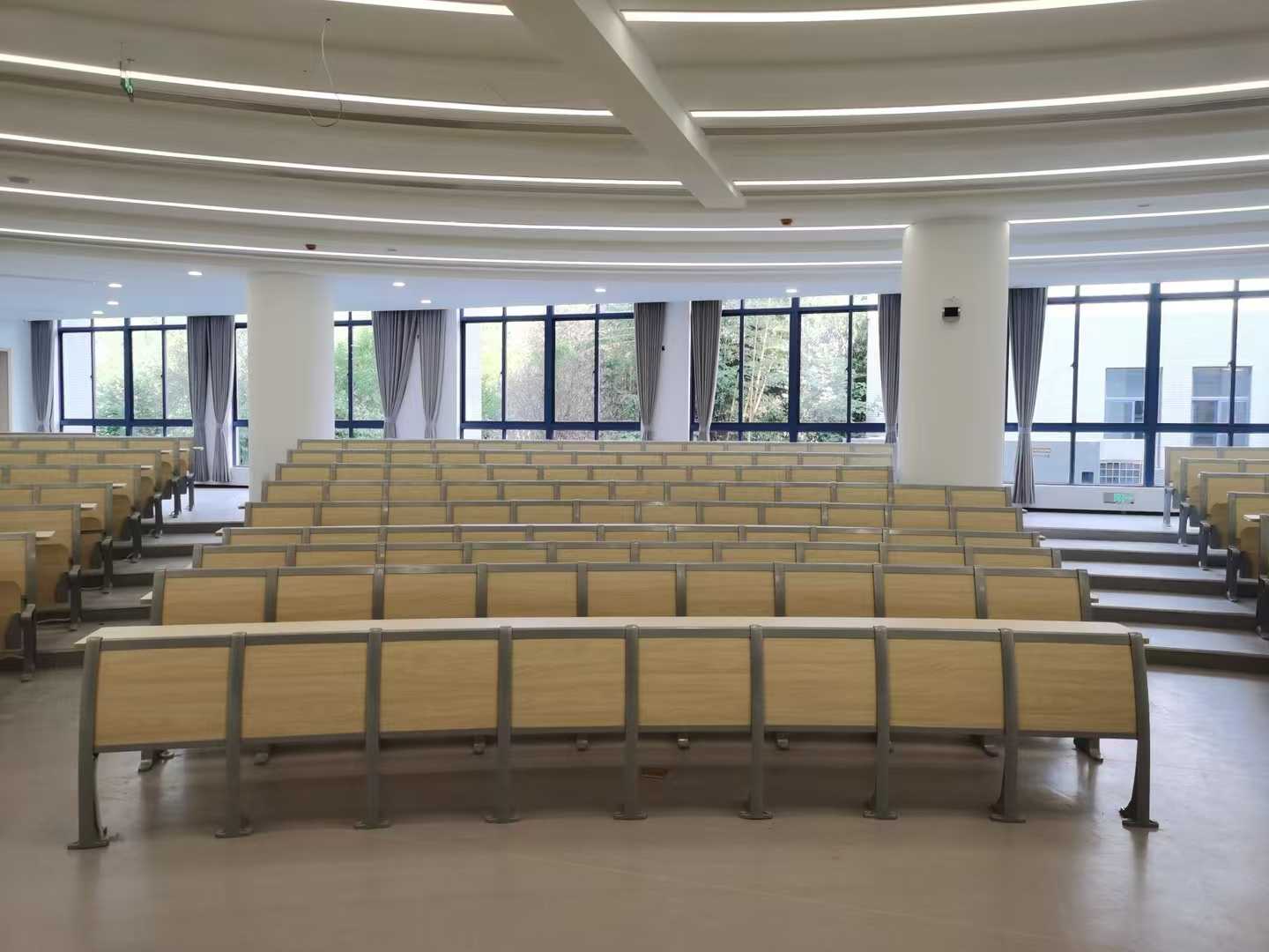 Empty lecture hall with light wood desks and chairs arranged in rows facing large windows.