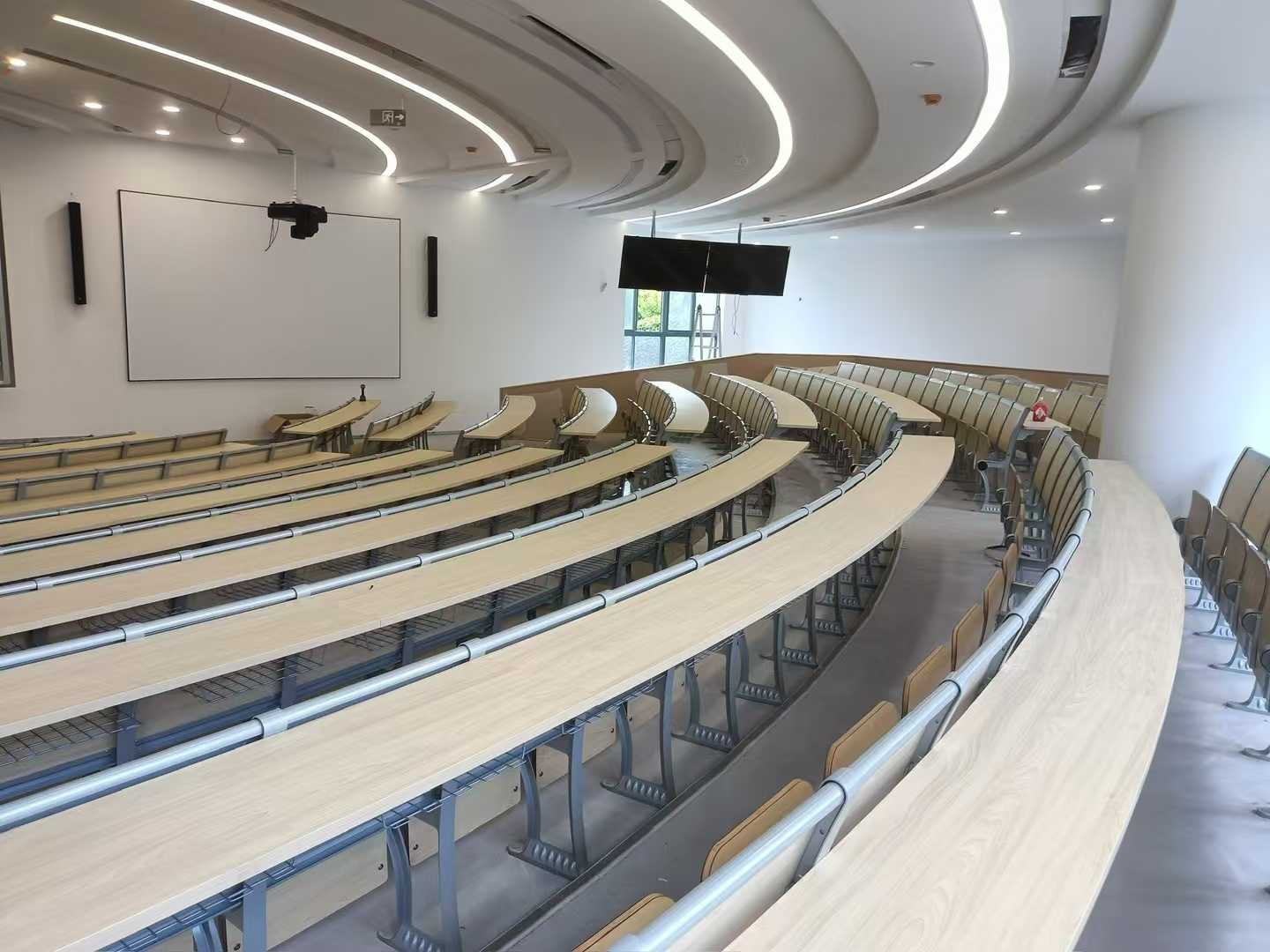 Empty curved lecture hall with light wood desks and chairs, a large projection screen, and modern lighting.