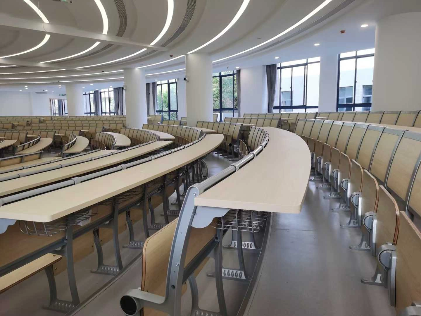 Modern lecture hall with curved rows of light beige desks and chairs.