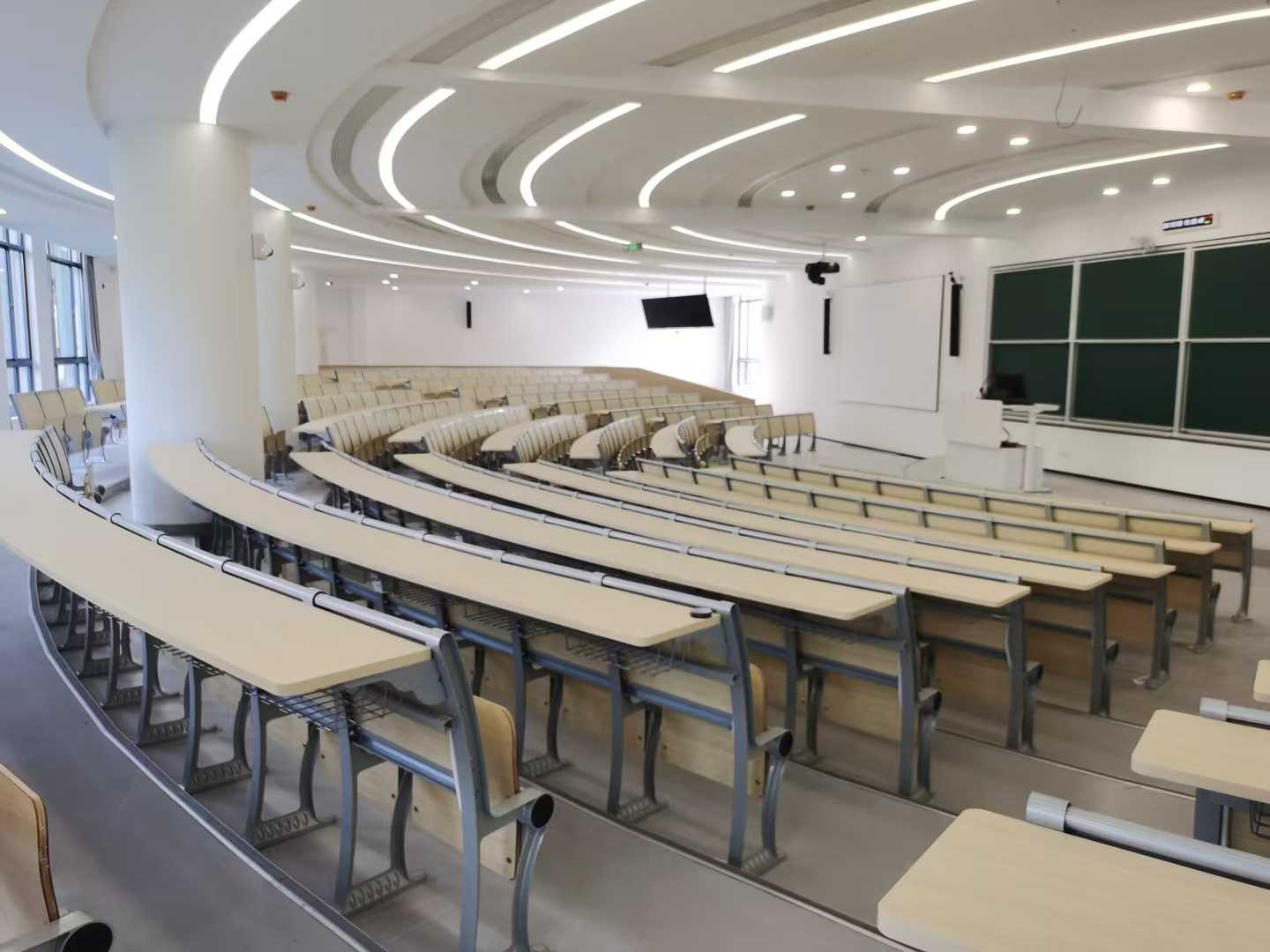 Modern lecture hall with curved rows of light wood desks and chairs, a large whiteboard, and a projector screen.