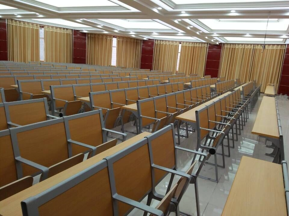 Lecture-hall-with-rows-of-light-brown-wooden-desks-and-chairs. A lecture hall filled with rows of light wood student desks and chairs.