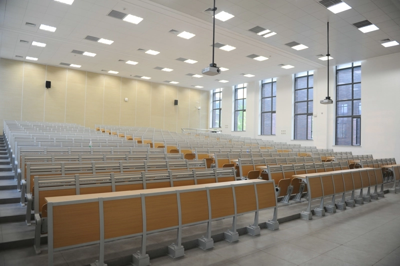 customizable-educational-seating Empty lecture hall with rows of light wood desks and chairs, a projector hanging from the ceiling, and large windows letting in natural light.
