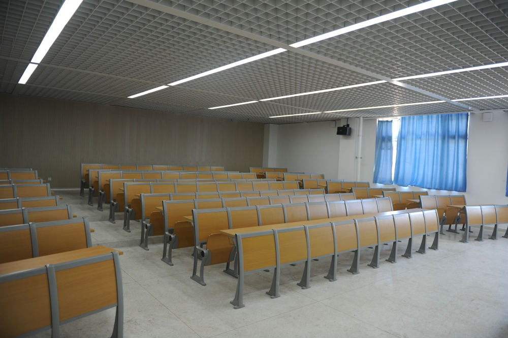 Empty classroom with light wood desks and chairs arranged in rows, under a grid ceiling with bright fluorescent lights. Empty classroom with light wood desks and chairs arranged in rows, under a grid ceiling with bright fluorescent lights.