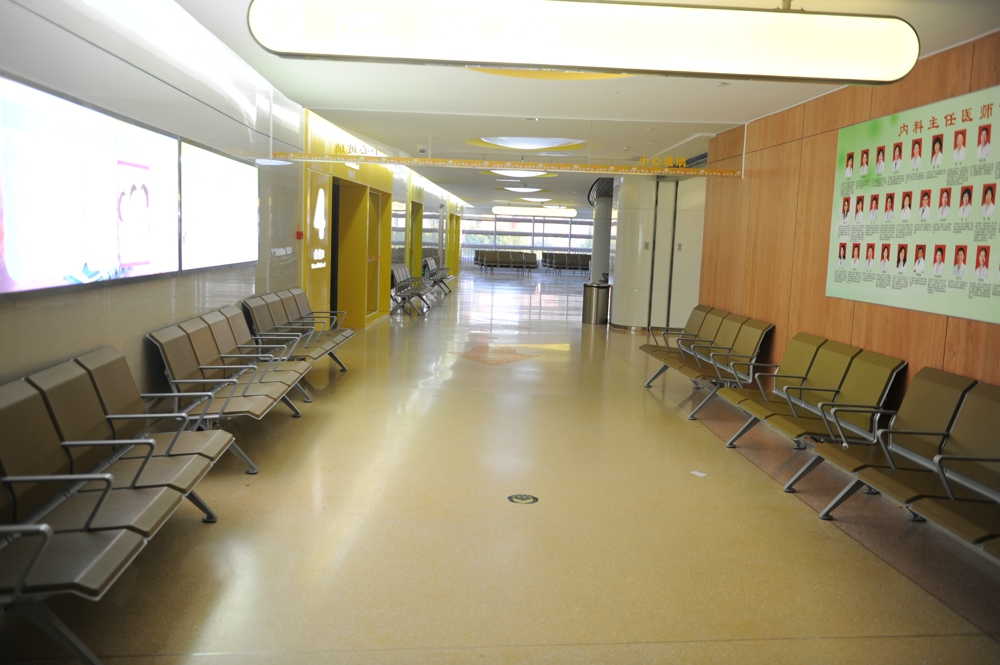 Empty waiting area in a hospital with rows of beige chairs facing each other, and a wall displaying portraits of medical staff. Empty waiting area in a hospital with rows of beige chairs facing each other, and a wall displaying portraits of medical staff.