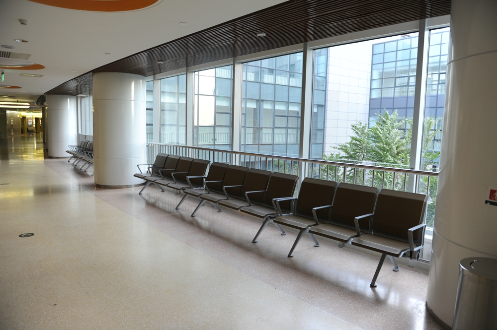 Empty waiting area in a modern building with a row of metal and upholstered chairs facing large windows overlooking city buildings and trees. Empty waiting area in a modern building with a row of metal and upholstered chairs facing large windows overlooking city buildings and trees.