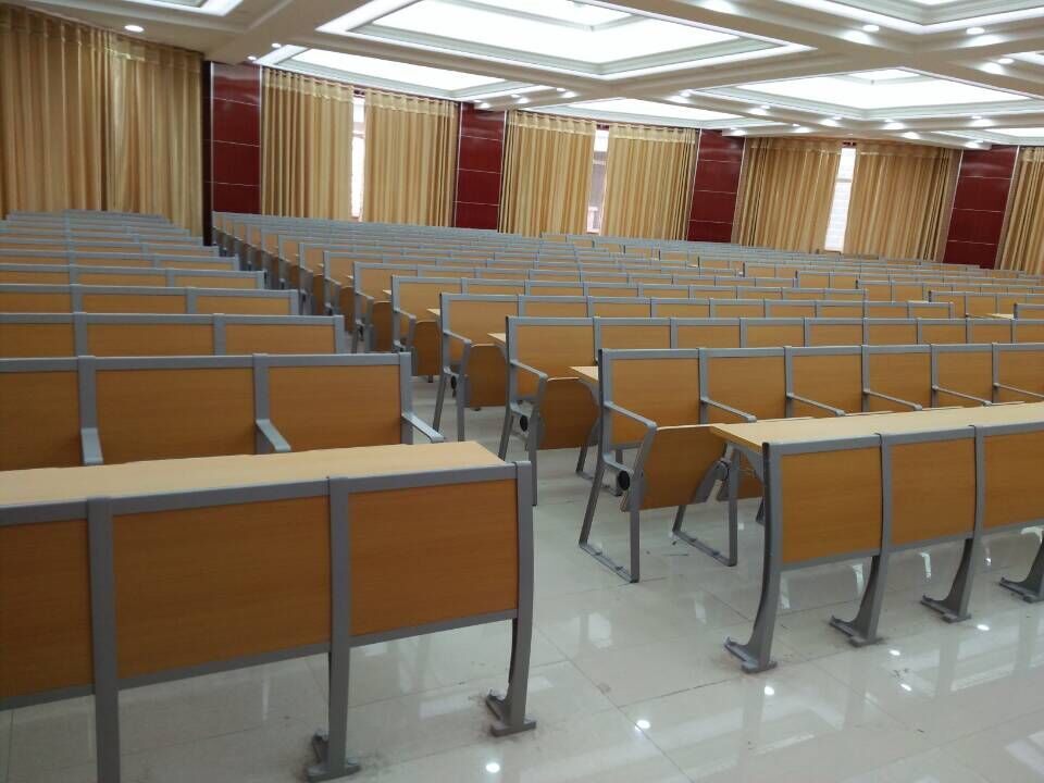 Rows of light brown wooden lecture hall desks and chairs in a large room with beige curtains. Rows of light brown wooden lecture hall desks and chairs in a large room with beige curtains.