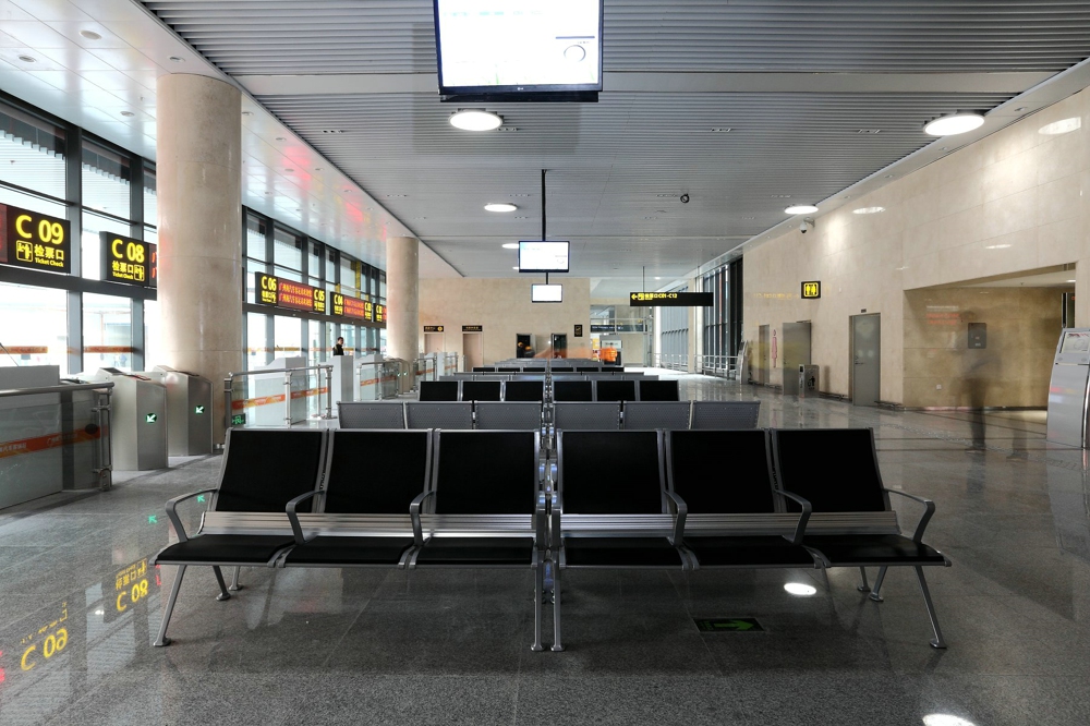 Empty waiting area with rows of metal and black cushioned chairs, digital displays on the ceiling and walls, and ticket counters visible in the background. Empty waiting area with rows of metal and black cushioned chairs, digital displays on the ceiling and walls, and ticket counters visible in the background.