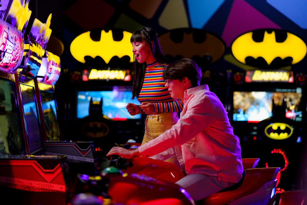 Excited young couple playing a motorcycle racing game in a brightly lit arcade featuring Batman games.