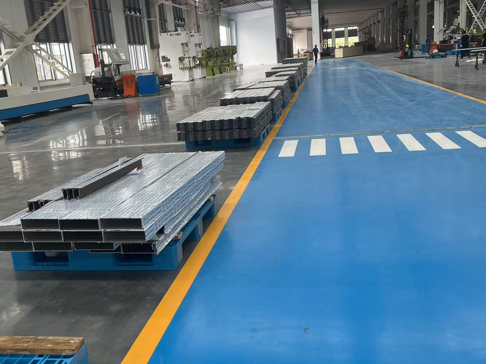 Stacked metal profiles on pallets in a factory with a blue floor walkway.